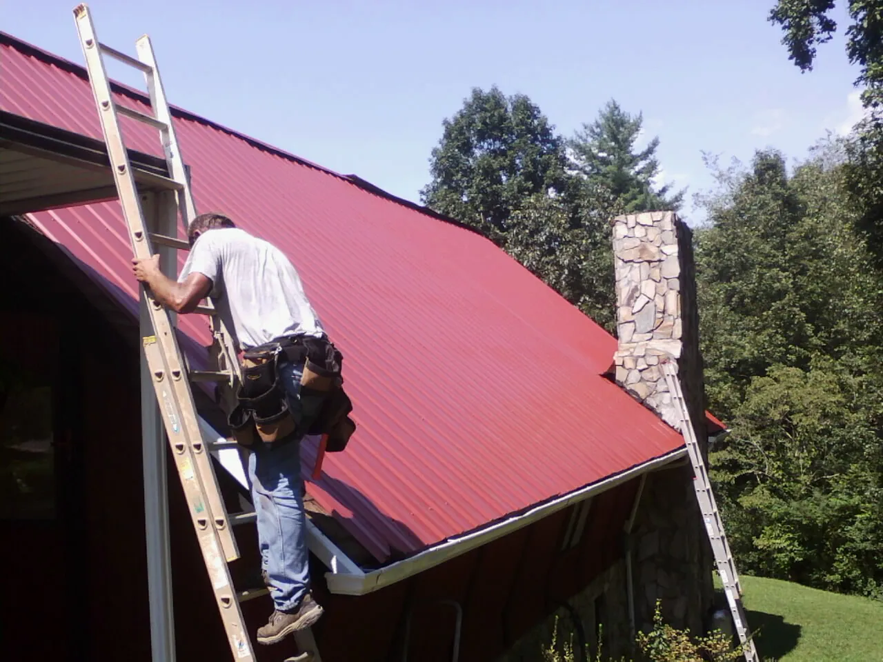Roofer on a ladder beside a red metal roof and stone chimney — Soffit & Fascia Repair by Roofing Brothers in Fort Mill